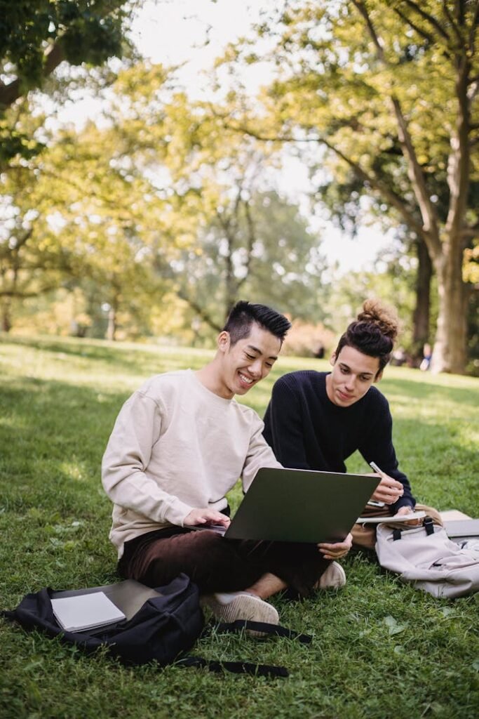 Full length content ethnic male students in casual outfits browsing laptop while sitting on meadow with copybooks and preparing for exam in green park